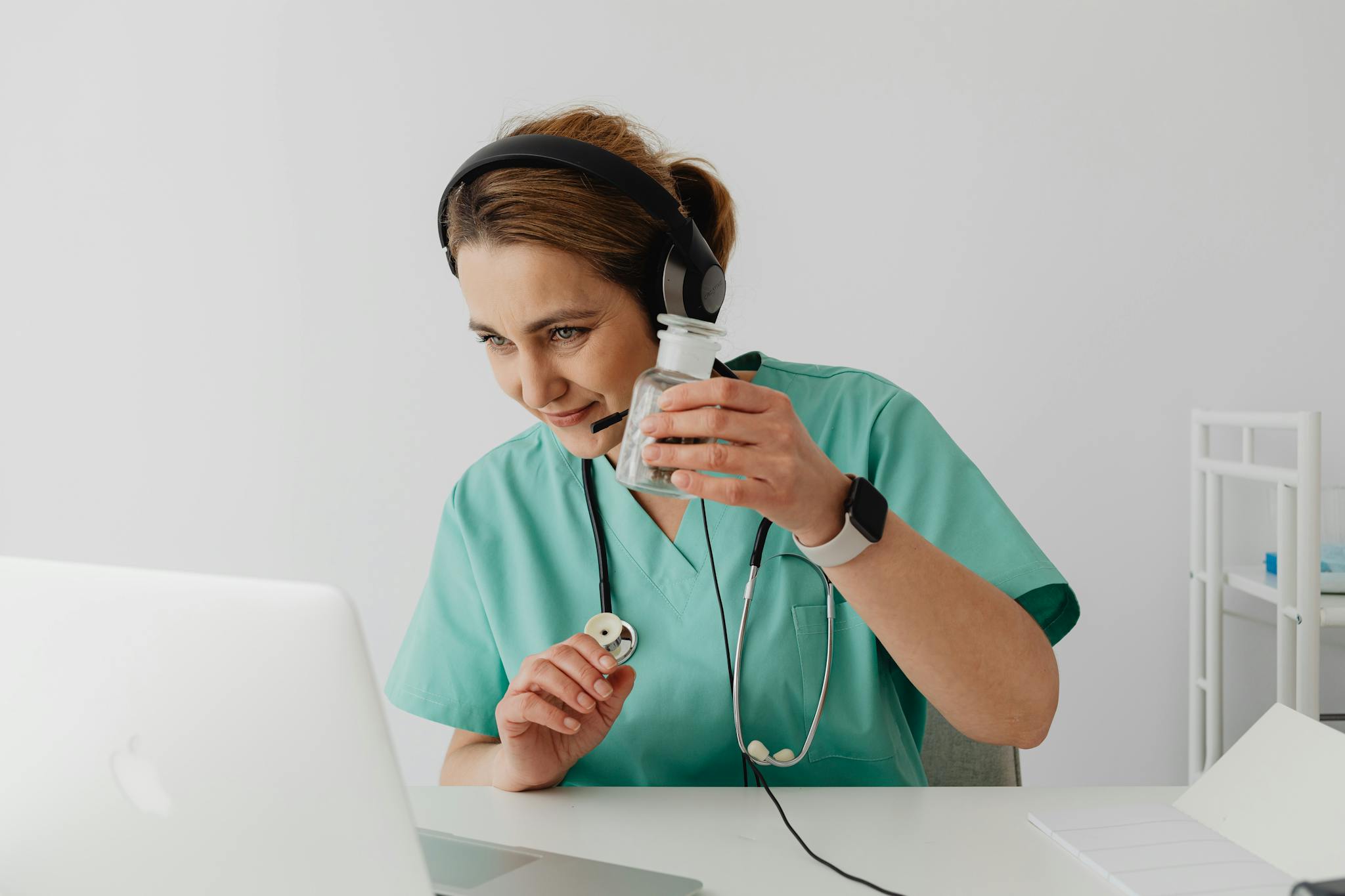 Doctor in scrubs having an online consultation using a laptop, holding pill bottle and smiling.