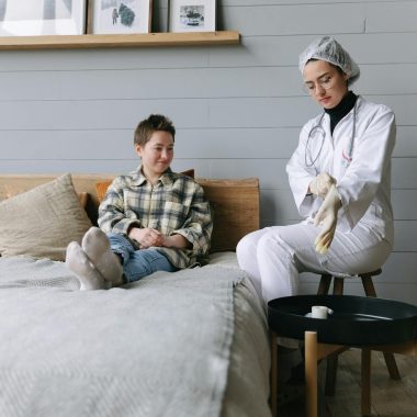 Patient sits on bed interacting with a doctor in a cozy bedroom environment.