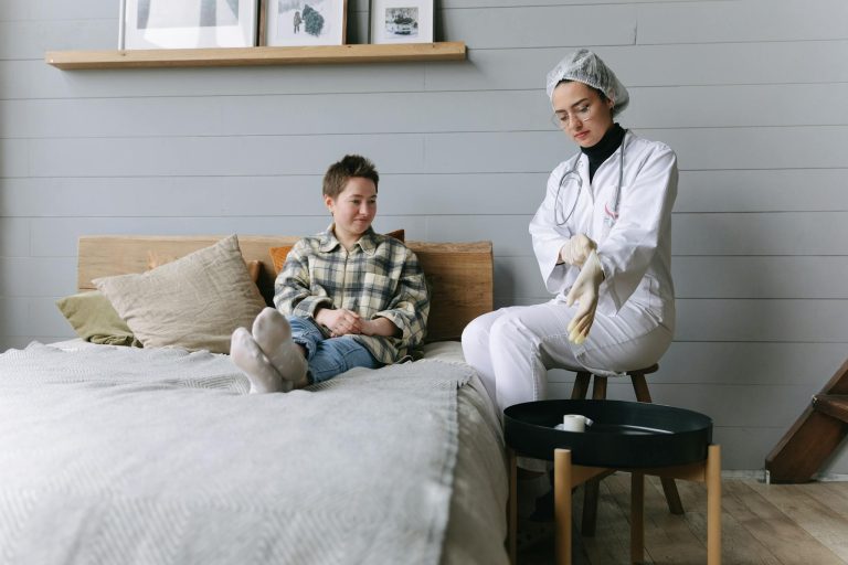 Patient sits on bed interacting with a doctor in a cozy bedroom environment.