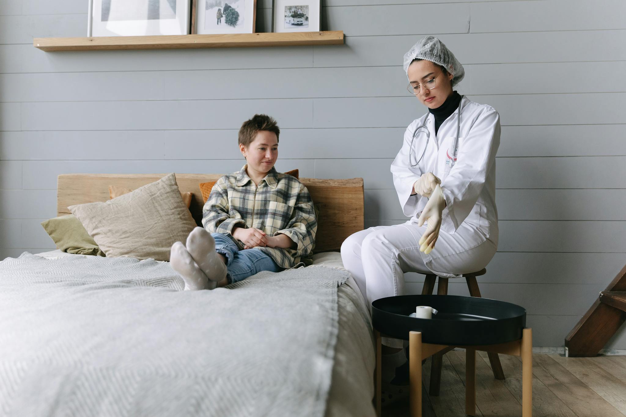 Patient sits on bed interacting with a doctor in a cozy bedroom environment.